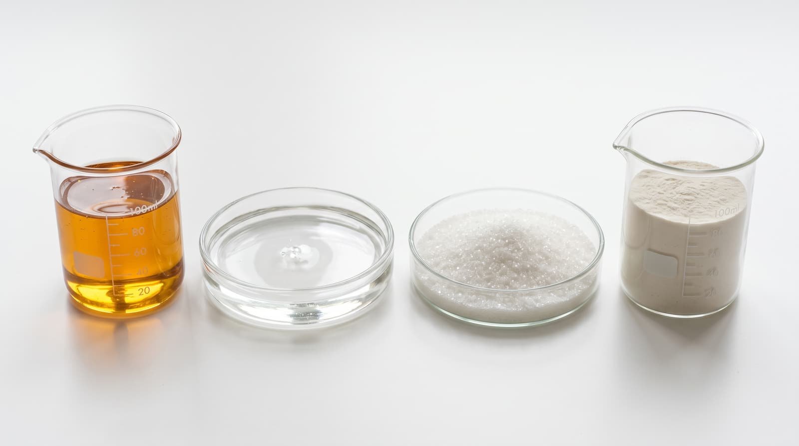 Glass beakers and dishes holding golden syrup, clear syrup, crystalline sweetener and fine powder on a white surface.